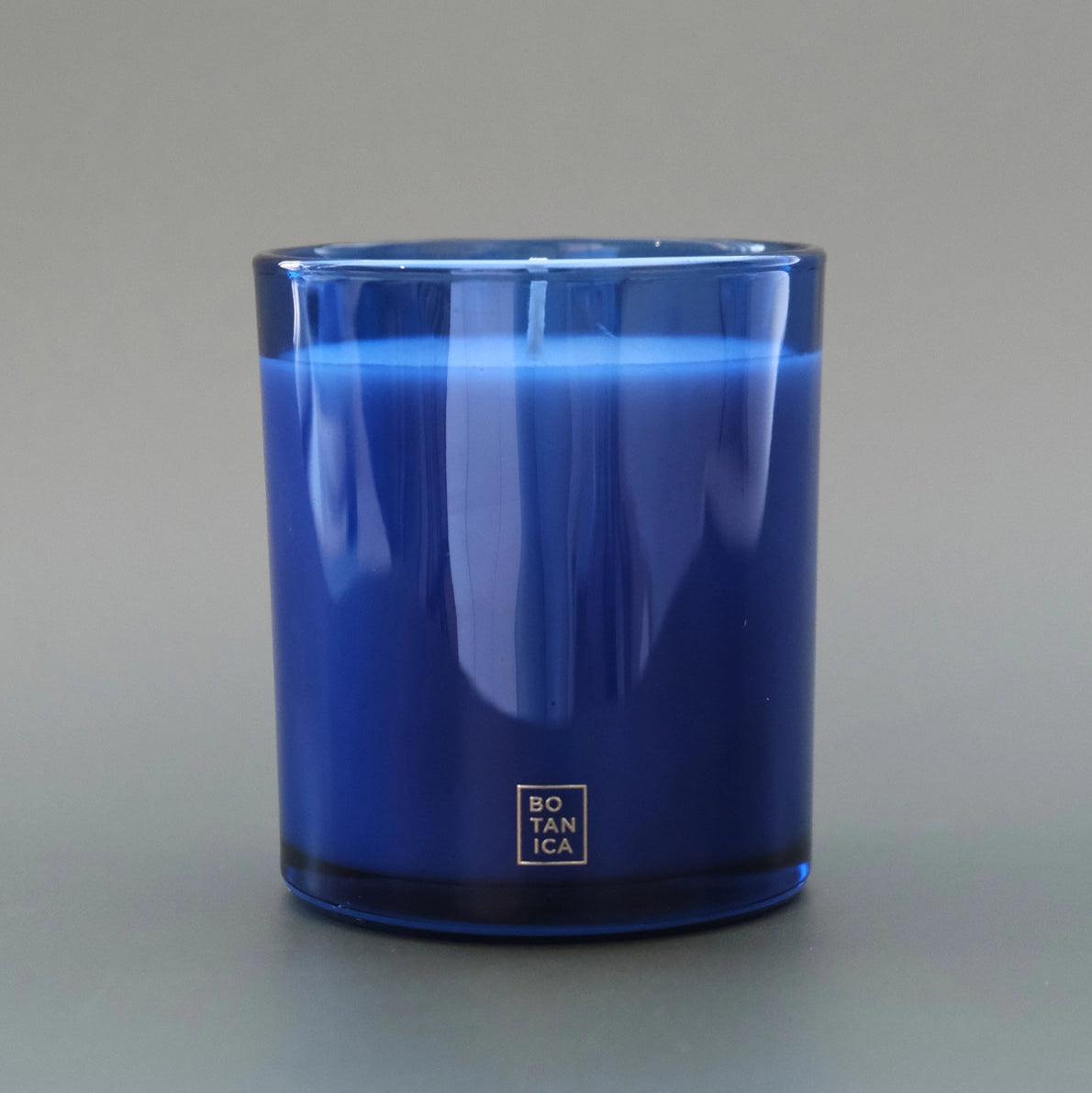 A cobalt blue glass candle jar with a silver square logo at the bottom, photographed against a neutral gray background.