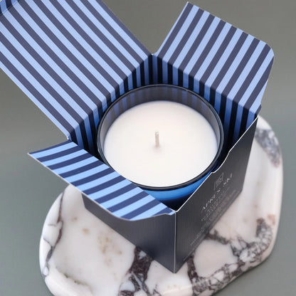 A white wax candle in a dark blue jar sits inside an open striped box with blue and white vertical bands, placed on a white and gray marble coaster against a smooth gray background.