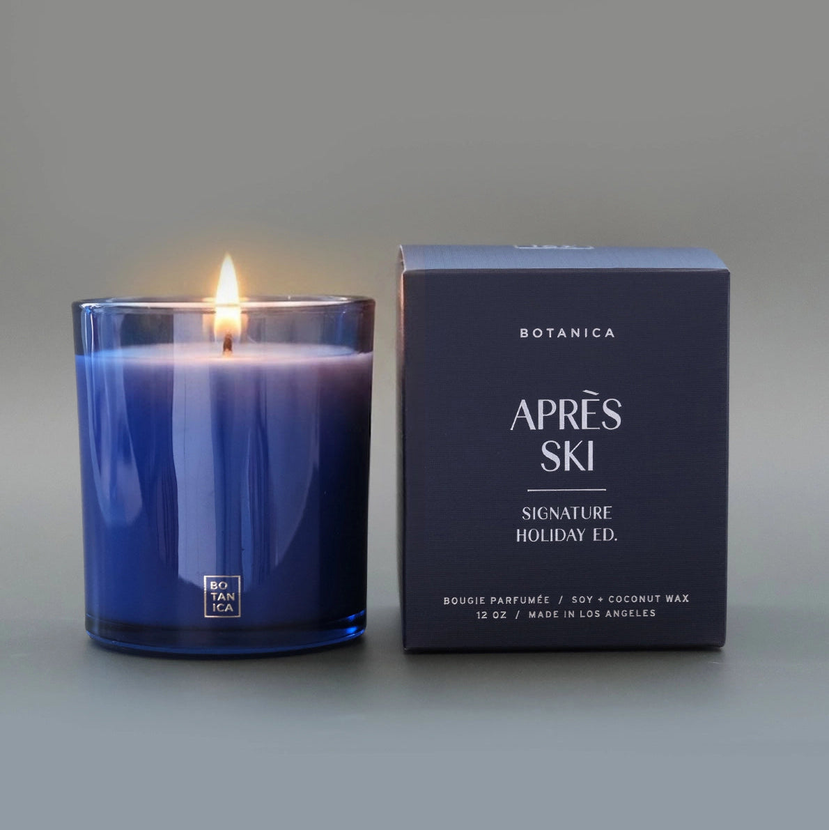 A lit candle in a blue glass jar next to a blue box embossed with silver text.