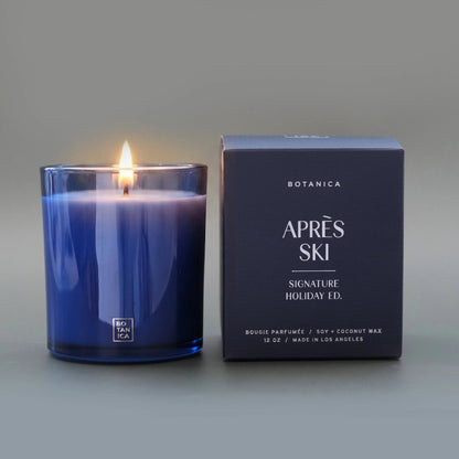 A lit candle in a blue glass jar next to a blue box embossed with silver text.