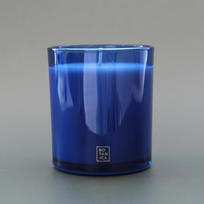 A cobalt blue glass candle jar with a silver square logo at the bottom, photographed against a neutral gray background.