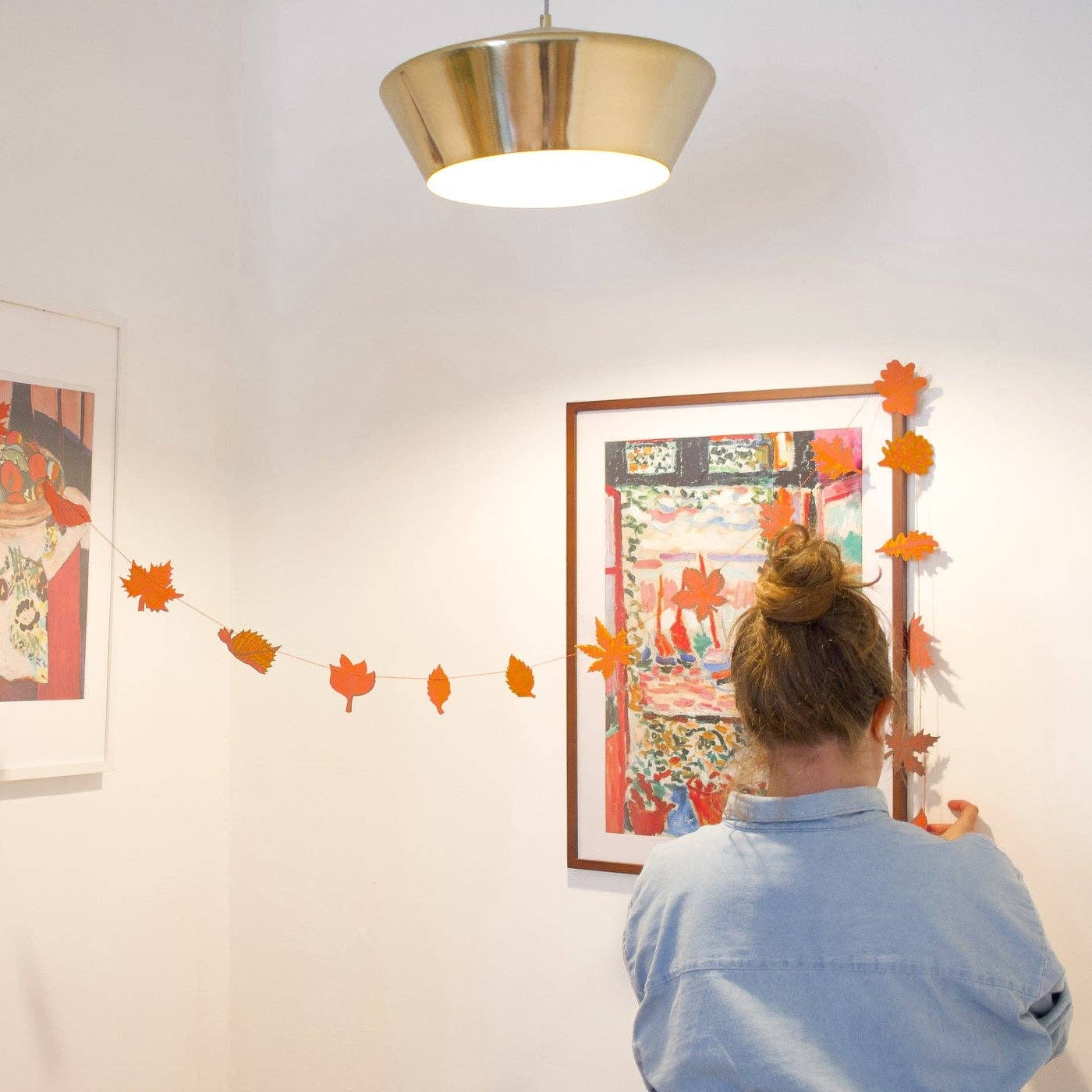 A woman in a light blue shirt is seen from behind, hanging orange paper leaves along a string between two framed art prints on a white wall.  