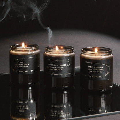 Three dark grey glass jar candles with black labels are lit and displayed on a reflective black surface, with smoke drifting above them against a dark background.