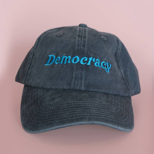 A washed dark blue baseball cap with the word 'Democracy' embroidered in bright blue script across the front, set against a solid pink background.