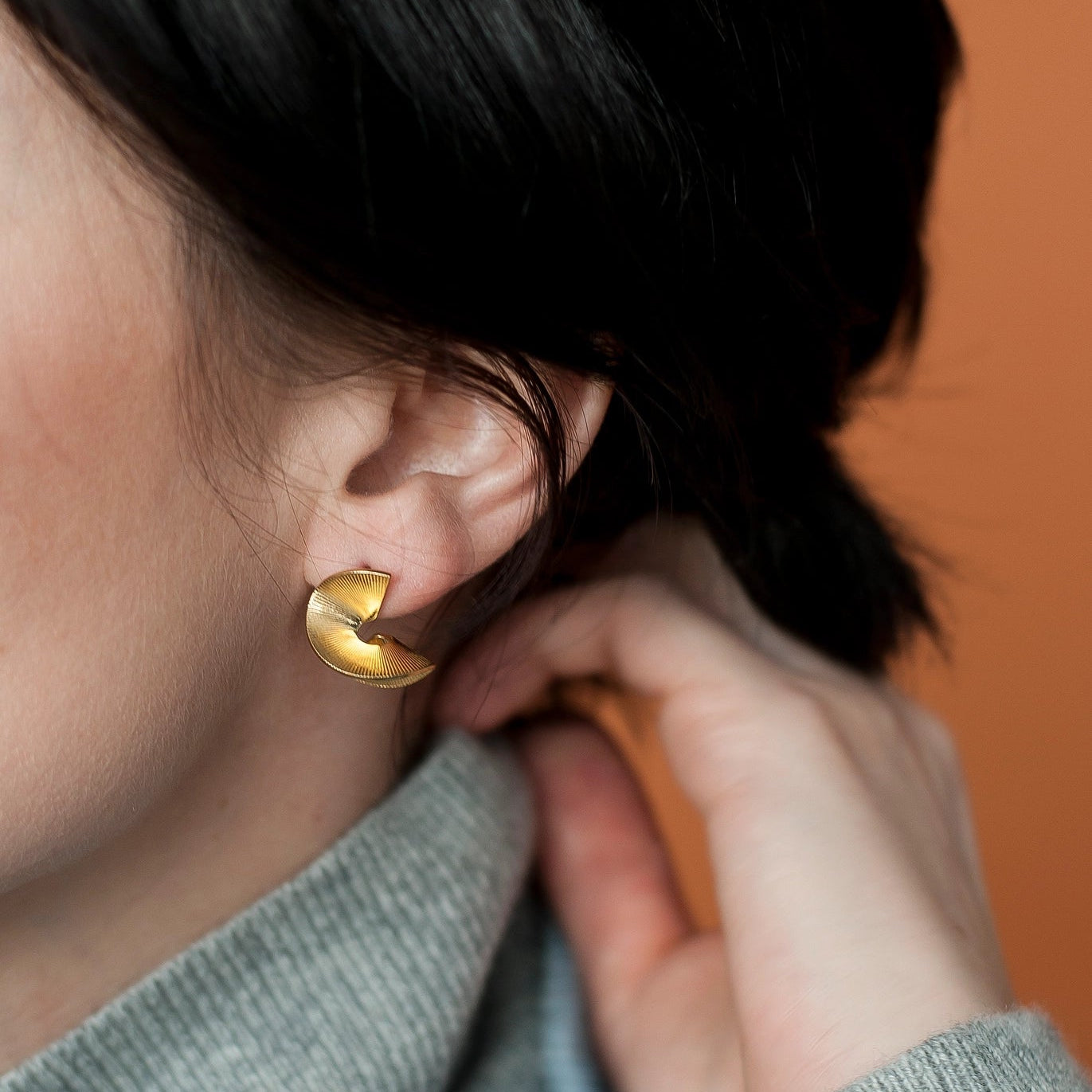 Person adjusting a gold earring on their ear against an orange background