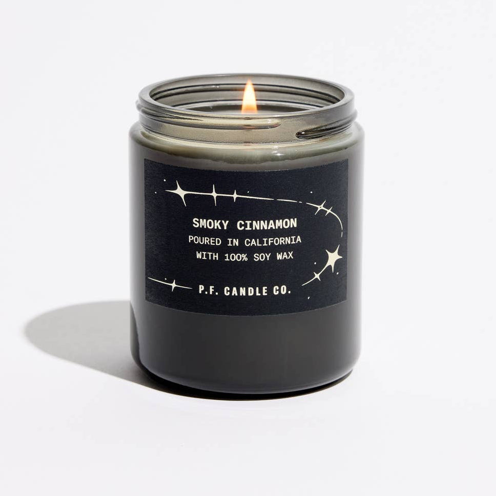 A lit candle in a dark glass jar with a black label reading 'SMOKY CINNAMON' and ‘P.F. Candle Co'