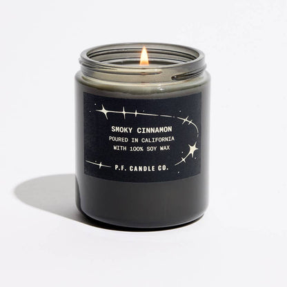 A lit candle in a dark glass jar with a black label reading 'SMOKY CINNAMON' and ‘P.F. Candle Co'