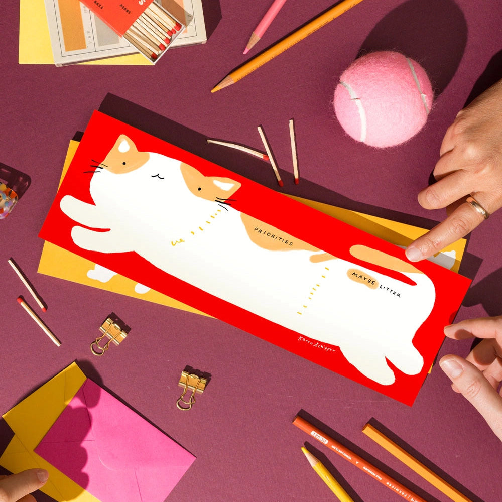 Red notepad with cat design held by hands on a purple surface with stationery items.