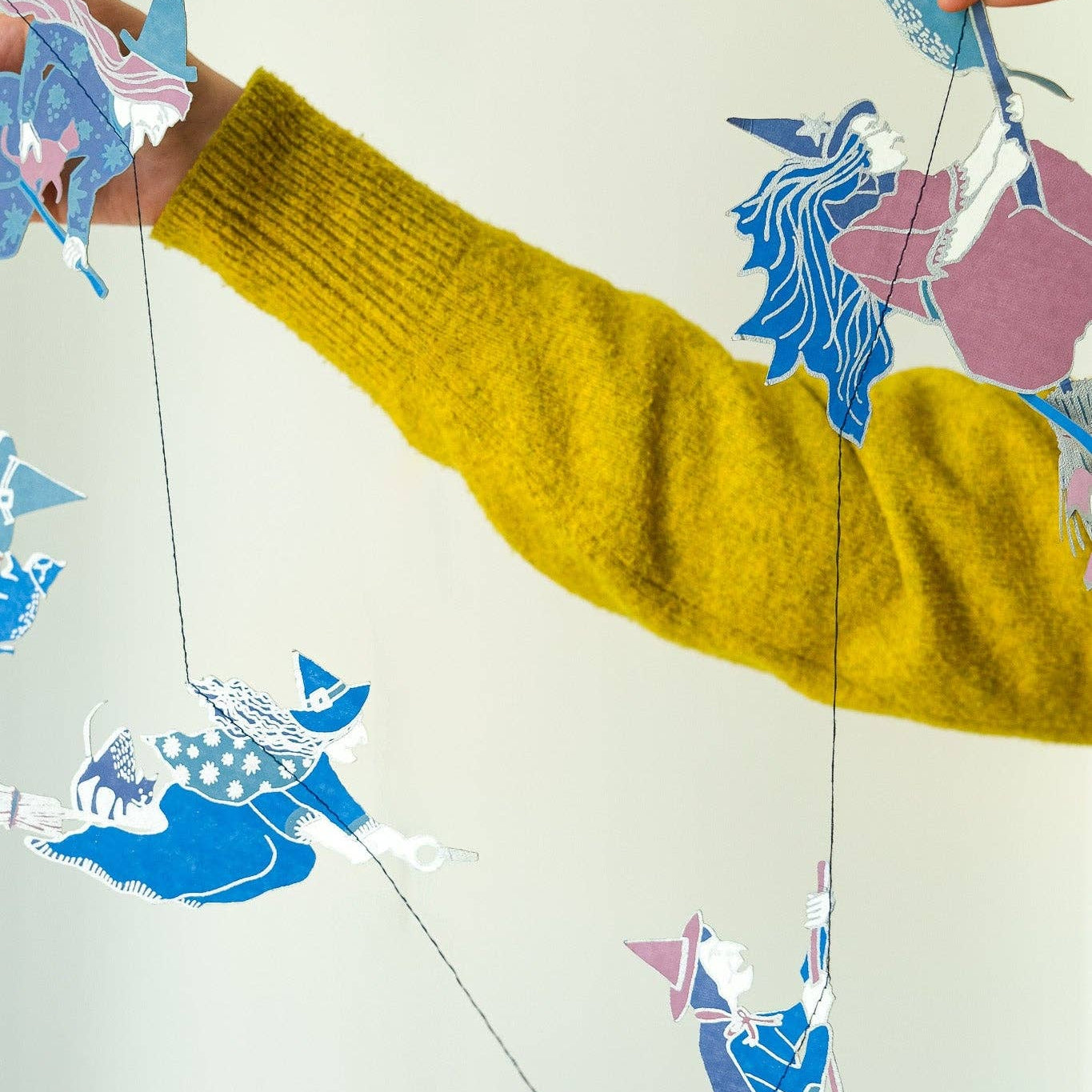 A person wearing a chunky mustard yellow knit sweater holds up a string of colorful paper witch cutout figures against a plain light background.