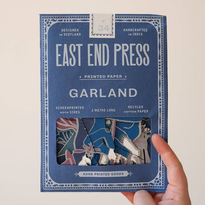 A hand holding a blue package of East End Press Garland printed paper against a plain light background. The package features white text and a window showing the colorful patterned sheets inside.