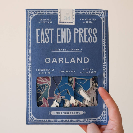 A hand holding a blue package of East End Press Garland printed paper against a plain light background. The package features white text and a window showing the colorful patterned sheets inside.